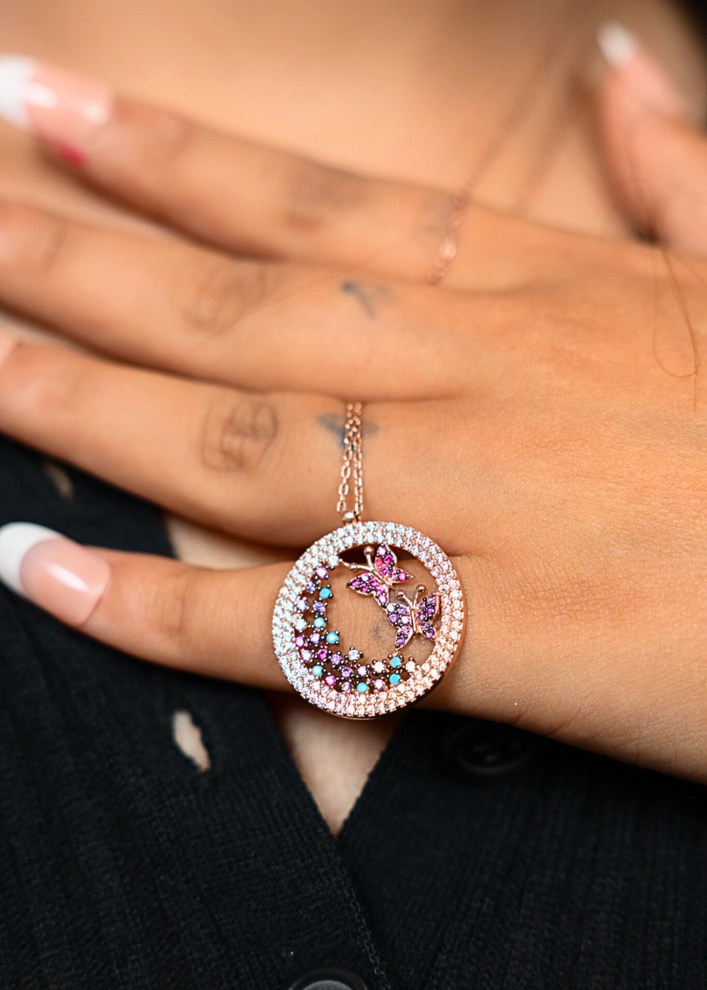 Close-up of a hand wearing a decorative necklace with a circular pendant featuring colorful stones and butterflies.